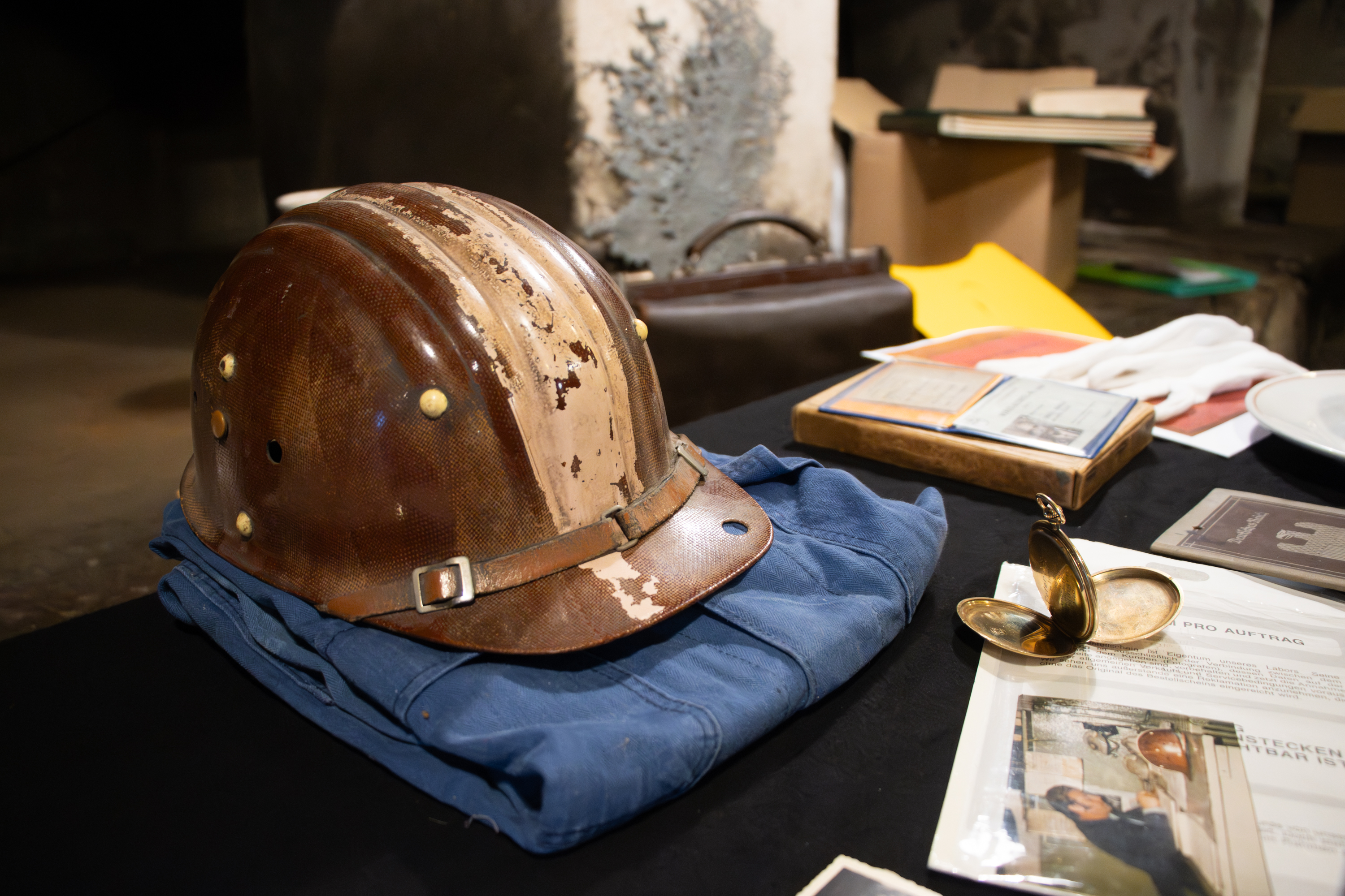 Mehrere Ausstellungsstücke liegen auf einem Tisch. Darunter befindet sich ein brauner Arbeitshelm, blaue Arbeitsklamotten und eine goldene Taschenuhr.