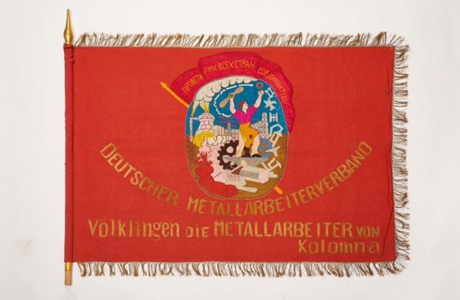 Eine rote sozialistische Flagge mit der Beschriftung: "Deutscher Metallarbeiterverband. Völklingen Die Metallarbeiter von Kolomna"